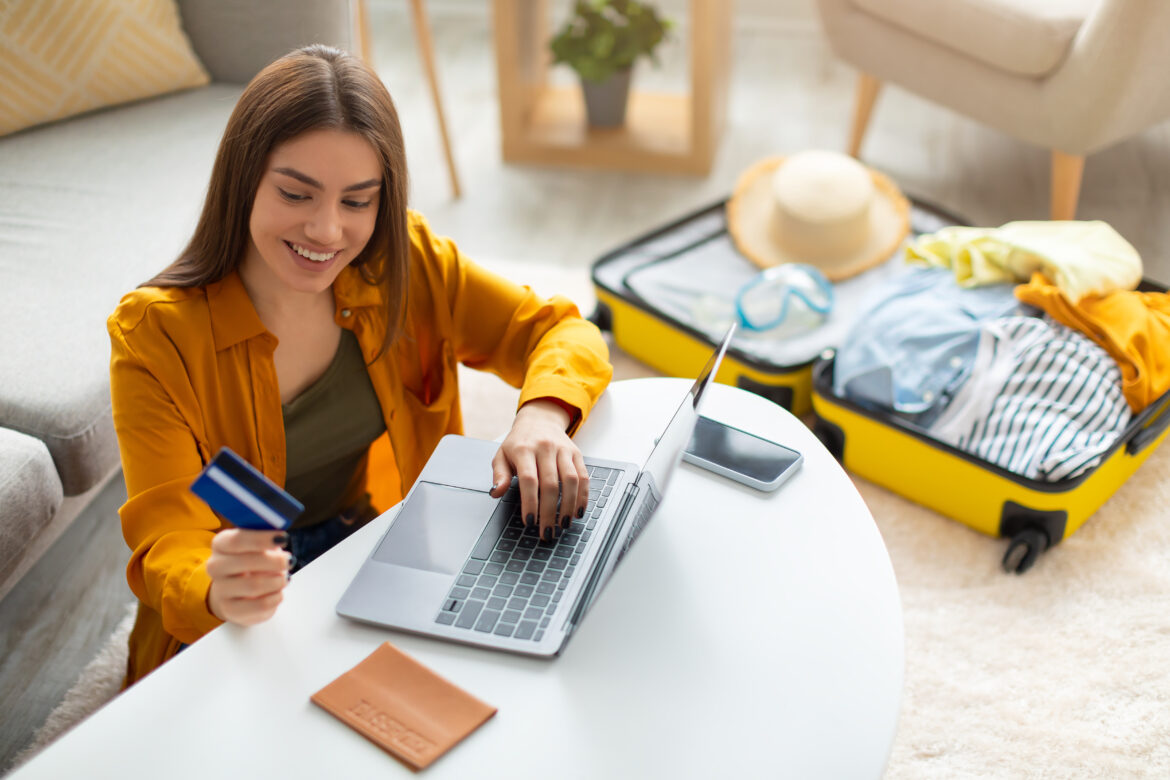Happy young woman with credit card booking vacation at travel agency or making hotel reservation online, using laptop computer, planning abroad trip at home, copy space