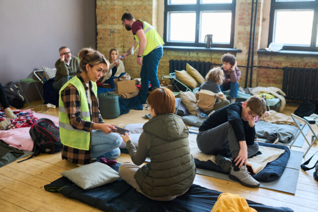 Young female volunteer passing smartphone to one of refugees sitting on mattress on the floor of spacious room serving as camp