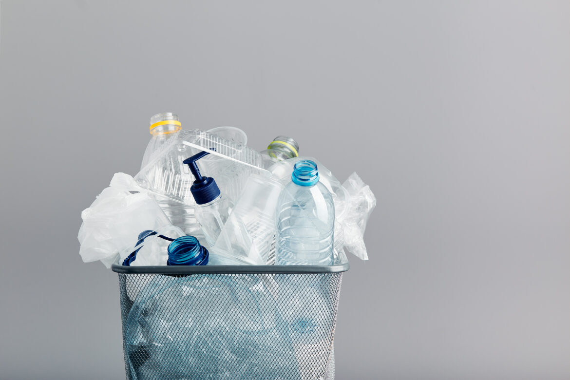 Heap of plastic bottles, cups, bags collected to recycling in a metal bin. Concept of plastic pollution and too many plastic waste. Trash with used plastic packagings over grey background with copy space at the top