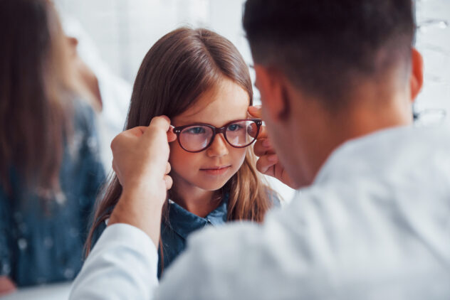 Young pediatrician in white coat helps to get new glasses for little girl.