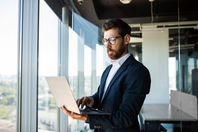 Successful middle aged businessman with laptop in hands standing in modern office interior near window, copy space. Serious male entrepreneur in formal suit working on computer