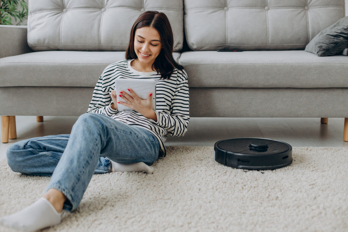 Woman sitting by the sofa using tablet while robot vacuum cleaner cleaning up the room