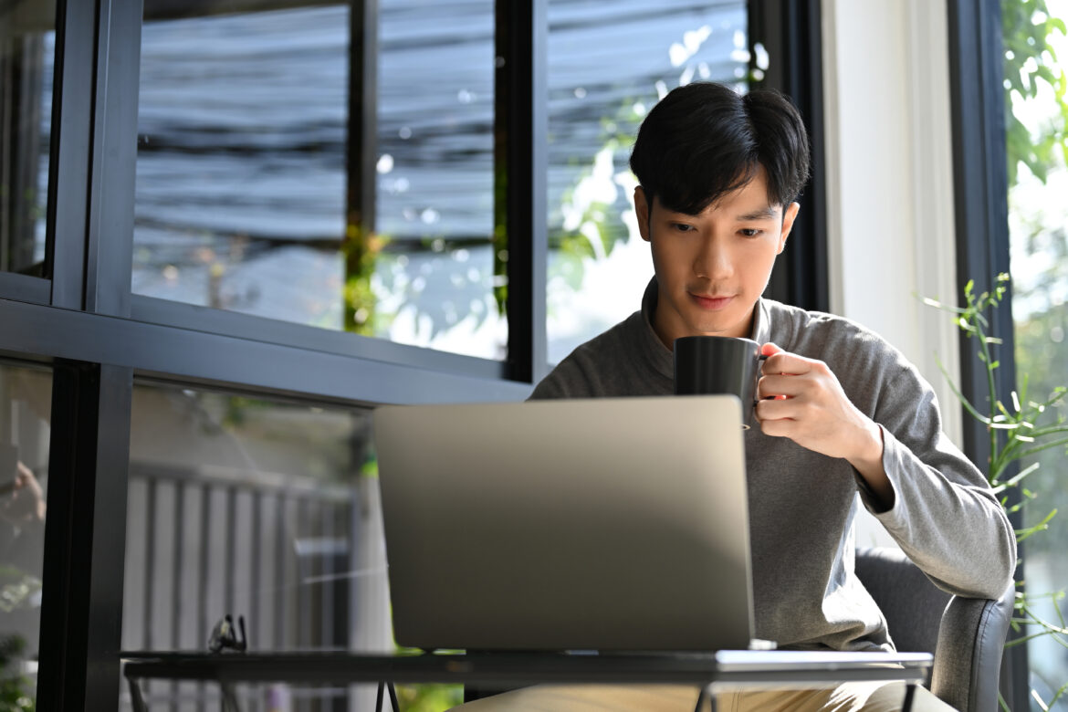 A young Asian man remote working at the coffee shop, sitting at the outdoor seat, sipping coffee while focusing on his work on laptop computer.
